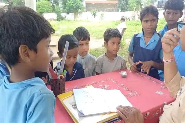 मेज पर खुली कॉपी के पास खड़े स्कूली बच्चों से बातचीत करता एक व्यक्ति
