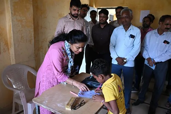एक स्कूल के कमरे में गुलाबी सूट पहने महिला अधिकारी मेज पर झुककर पीली टी-शर्ट पहने बच्चे को कॉपी पर लिखते हुए देख रही हैं, पीछे कई पुरुष अधिकारी खड़े हैं।