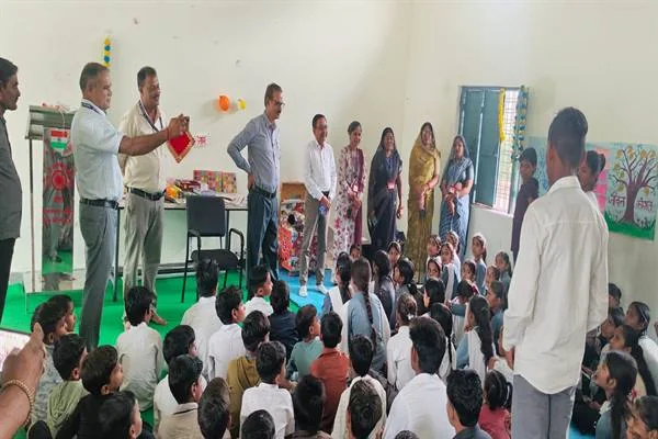 स्कूल के एक कमरे में फ़र्श पर बैठे छात्र-छात्राओं के सामने खड़े अधिकारी और शिक्षक-शिक्षिकाएँ, एक अधिकारी हाथ में लाल कपड़े में लिपटी वस्तु पकड़े हुए, पीछे दीवार पर पेड़ का चित्र बना बोर्ड।