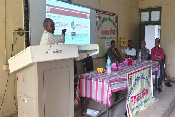 कक्षा-नुमा हॉल में एक वक्ता डायस के पास खड़े होकर प्रोजेक्टर स्क्रीन पर दिख रही हिंदी प्रस्तुति की ओर इशारा कर रहे हैं; दाईं ओर मेज के पीछे चार लोग बैठे हैं और मेज पर सेवा संकल्प अभियान लिखा बैनर लगा है।