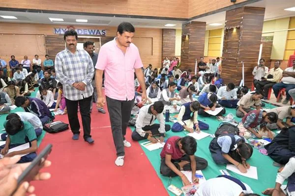 हॉल में हरी दरी पर बैठे स्कूली विद्यार्थी सफेद कागज पर चित्र बना रहे हैं, बीच के रास्ते से दो पुरुष गुजरते हुए, पीछे दर्शक कुर्सियों पर बैठे हैं।